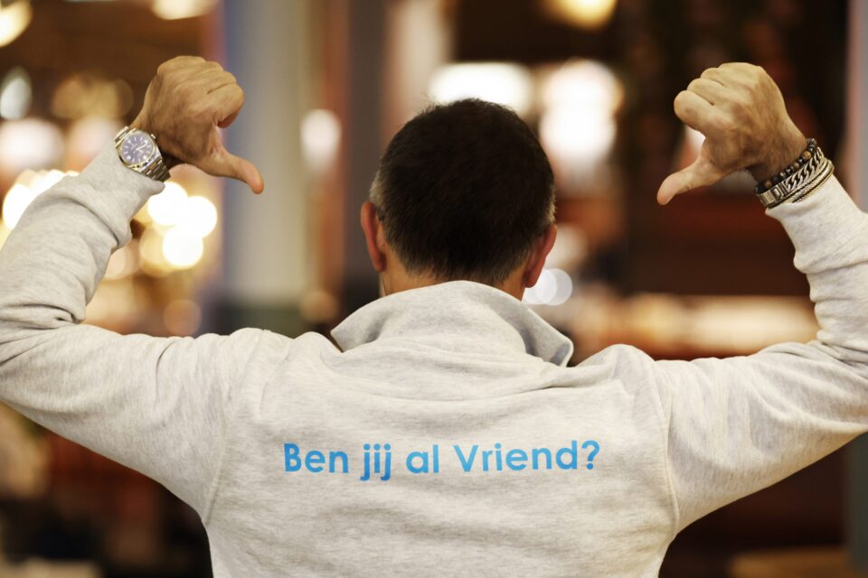 Achterkant van een meneer met een lichtgrijze trui en en met daarop de tekst 'Ben jij al Vriend?'.